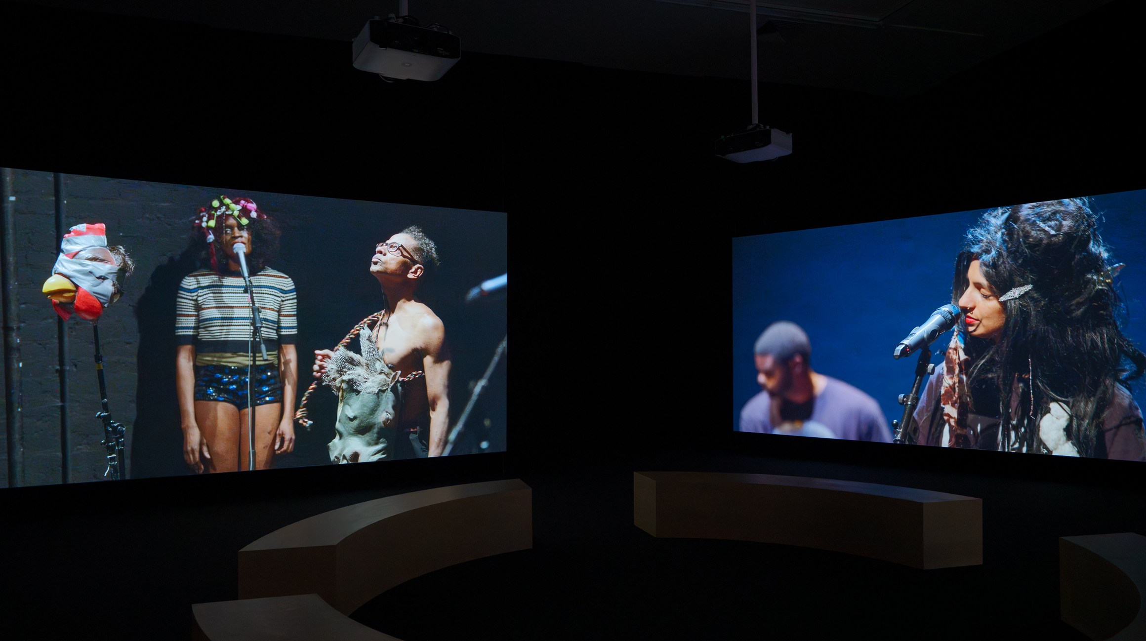 Two large screens display performers with microphones in a dark room, surrounded by curved seating and overhead projectors. From the Ralph Lemon retrospective at MoMA PS1.
