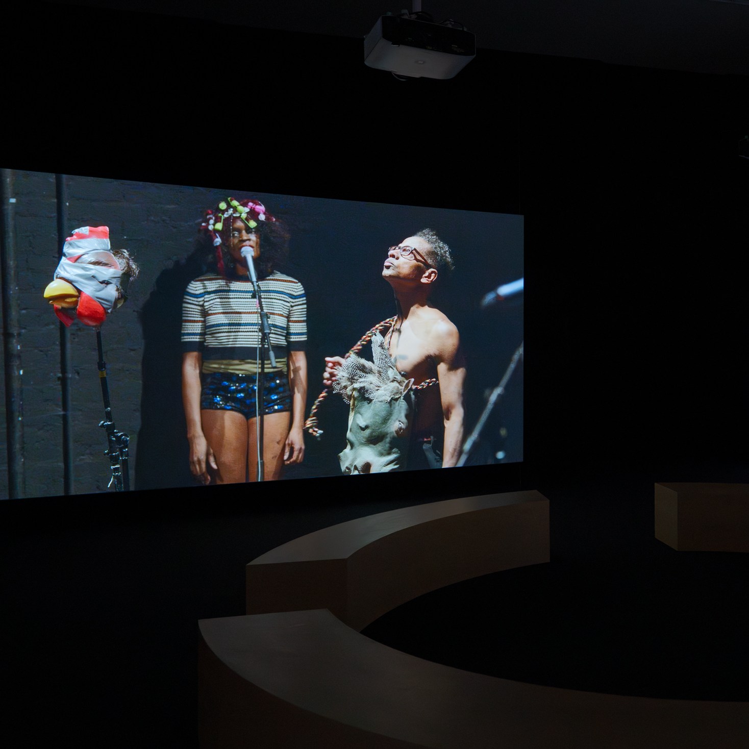 Two large screens display performers with microphones in a dark room, surrounded by curved seating and overhead projectors. From the Ralph Lemon retrospective at MoMA PS1.