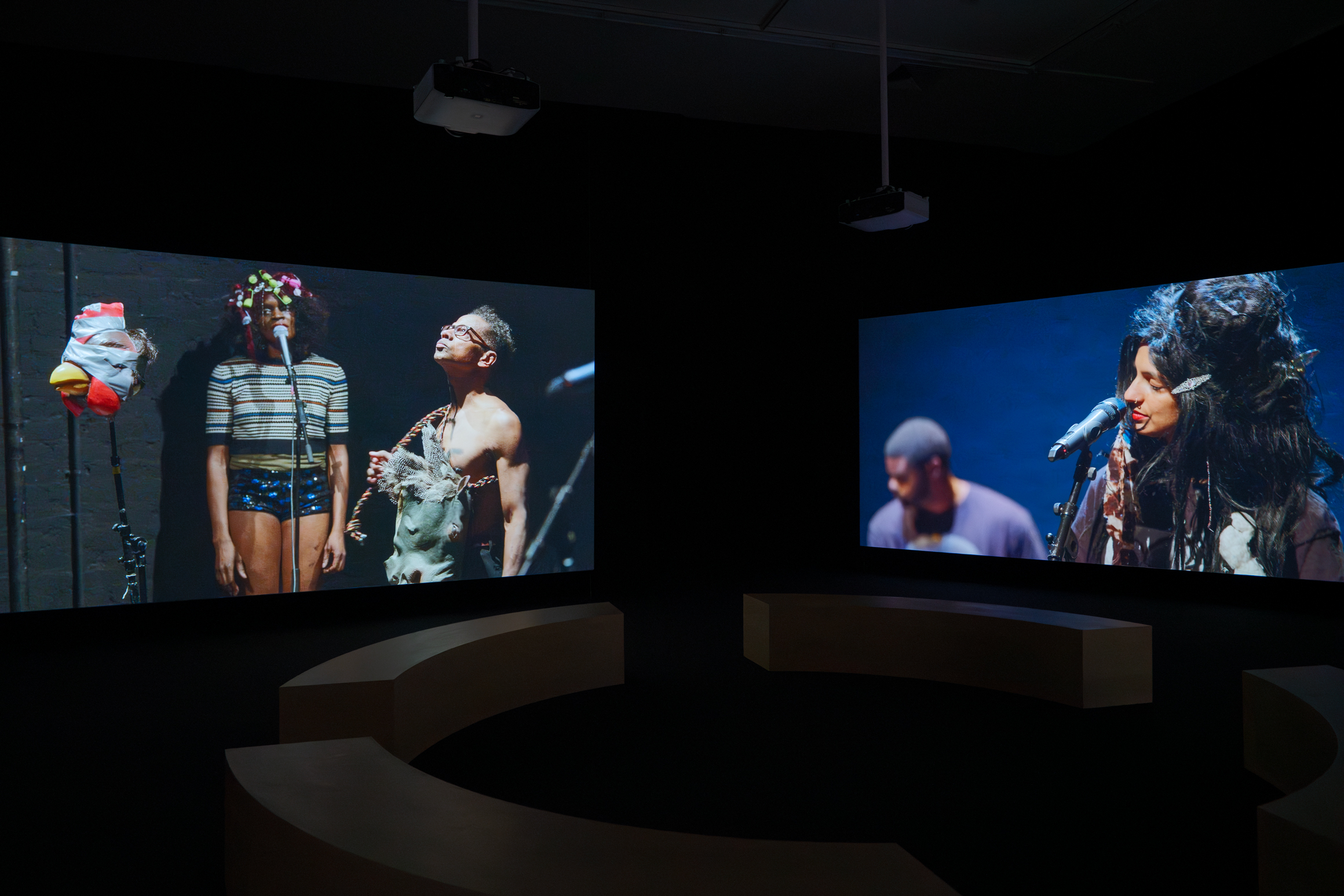 Two large screens display performers with microphones in a dark room, surrounded by curved seating and overhead projectors. From the Ralph Lemon retrospective at MoMA PS1.