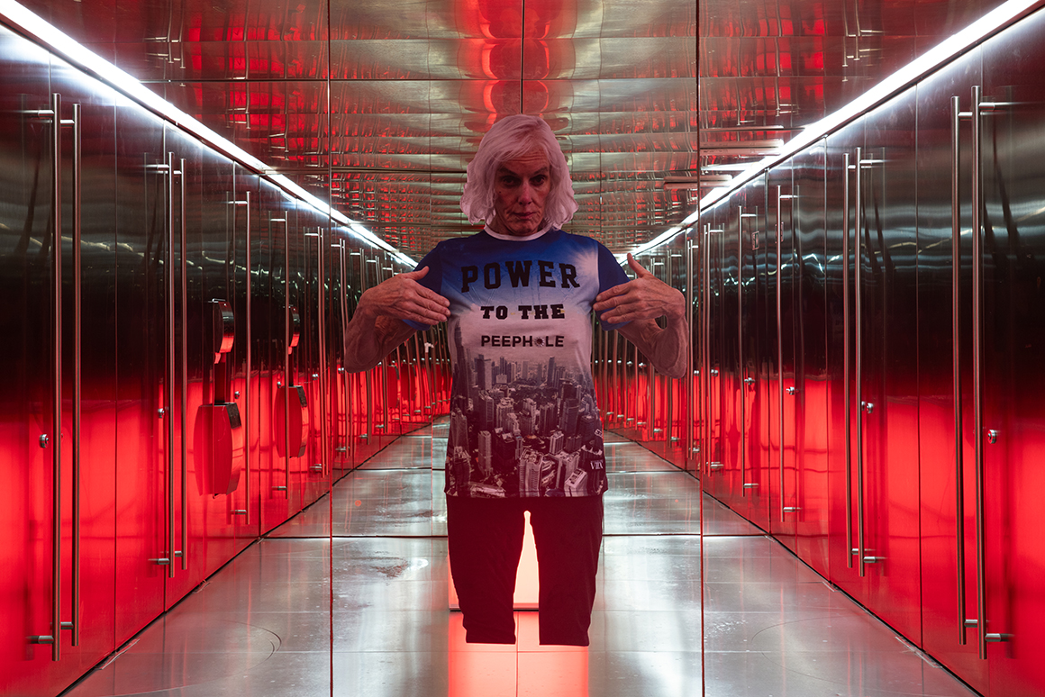 A decal of the artist Pippa Garner placed on a mirrored wall. Garner is wearing a shirt that says "POWER TO THE PEEPHOLE". The mirrored wall reflects a room with metal doors, ceiln, and flooring. There is white lighting above the doors, and a red light reflected on the bottom of the doors.
