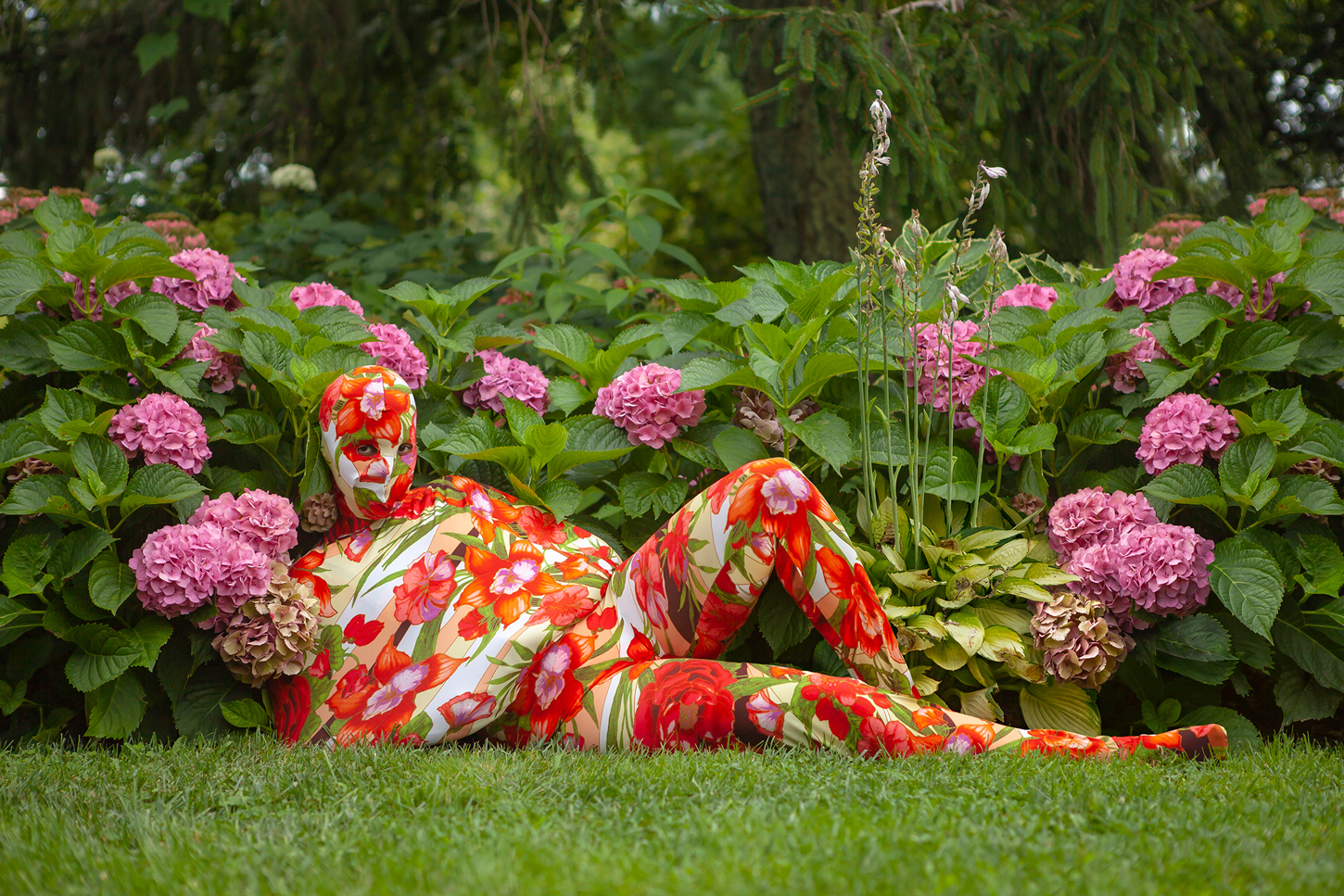 A person in a floral head-to-toe bodysuit reclines on grass. The bodysuit does not have individual sleeves for each arm, instead there is a sort of pocket or shelf covering both of the person's folded arms as one unit. The person is surrounded by pink hydrangea bushes with green leaves in a lush outdoor garden setting.