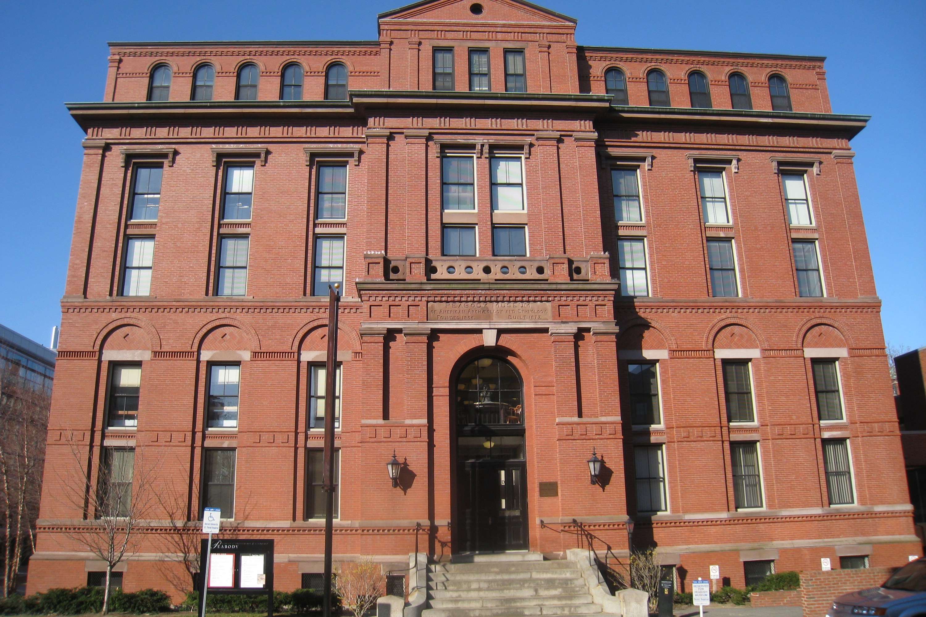 A large, red-brick building with a classic architectural style and arched windows. The sky above is clear and blue, and there is some greenery and signage in front of the building.