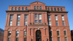 A large, red-brick building with a classic architectural style and arched windows. The sky above is clear and blue, and there is some greenery and signage in front of the building.
