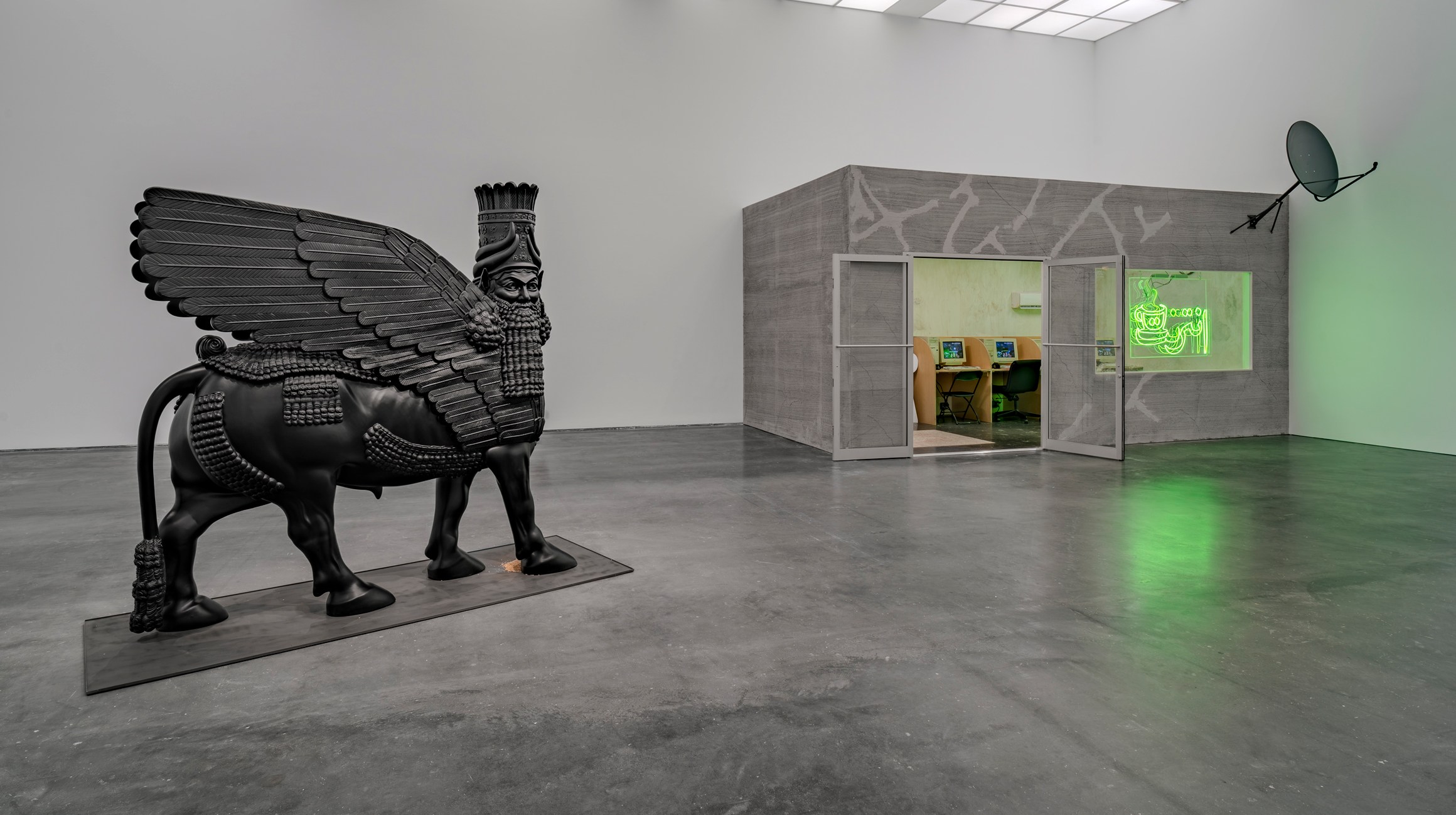 A black-winged bull sculpture stands on the left; a gray structure with doors, neon green signage, and workstations inside sits to the right. View of “Wafaa Bilal: Indulge Me,” 2025, Museum of Contemporary Art Chicago. From left: Lamassu (In a Grain of Wheat), 2025; In a Grain of Wheat: Cultivating Hybrid Futures in Ancient Seed DNA, 2021; Virtual Jihadi, 2008.