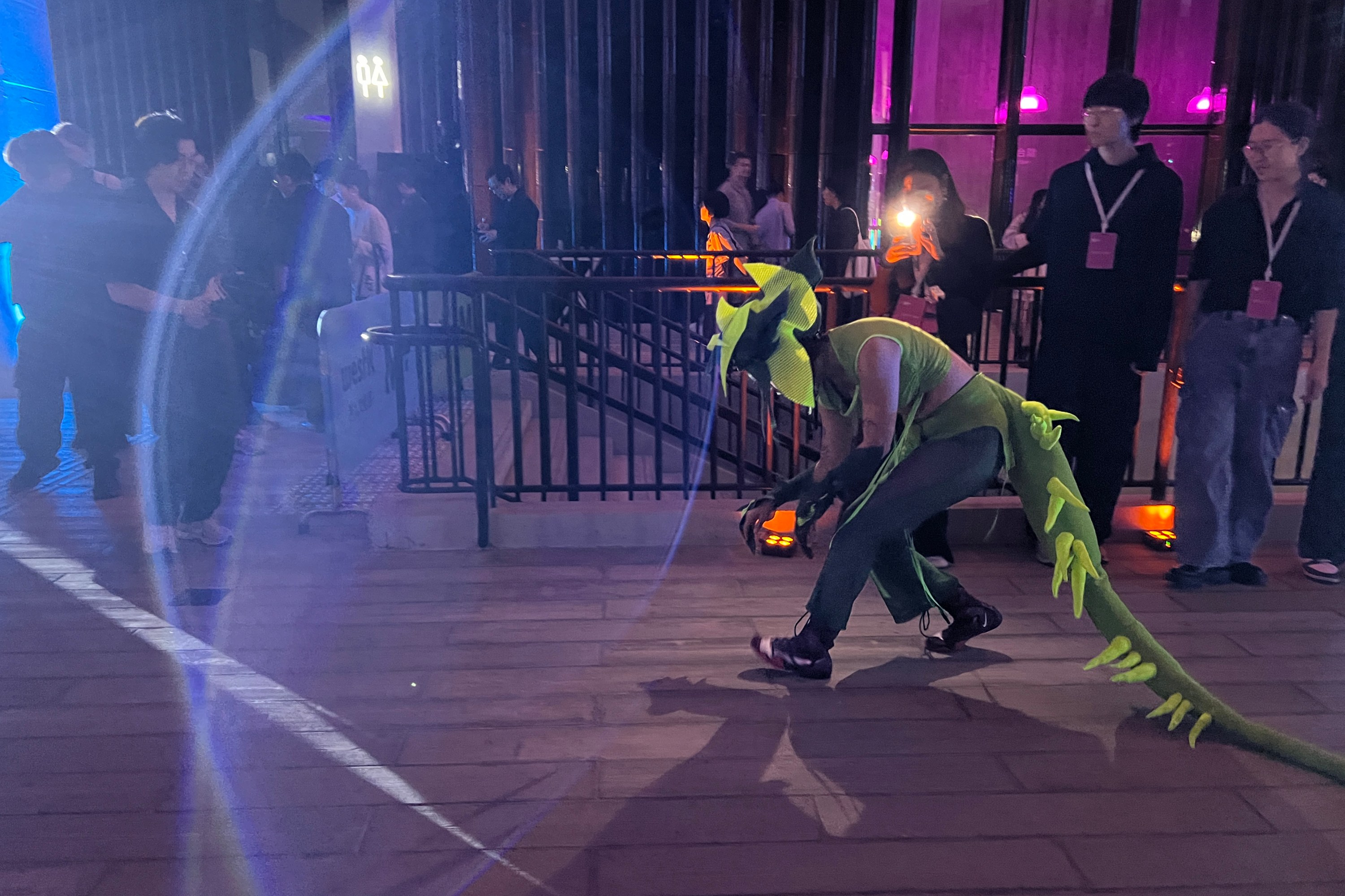 A performer dressed in a green reptilian costume with tail and horns crouches on a lit wooden floor near railing, surrounded by people and colorful lighting.