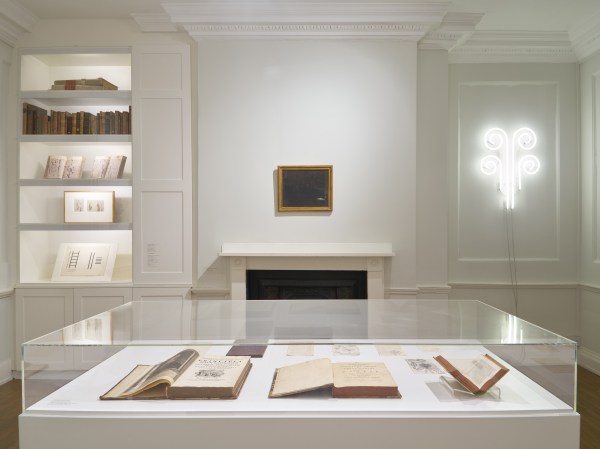Installation view of an art exhibition titled "Simon Moretti: Hereafter" at The Swedenborg Society. There is a glass vitrine in the foreground holding books. In the background is a book case, and artworks hung around a fireplace.