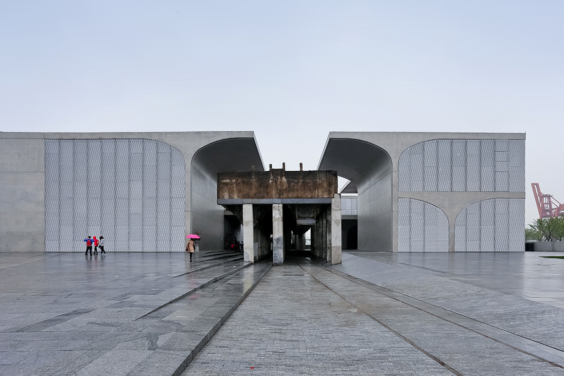 Atelier Deshaus, Long Museum West Bund. A modern concrete structure with large arch-shaped openings, rusted central beams, a tiled plaza, and people carrying umbrellas walking beneath a cloudy sky.