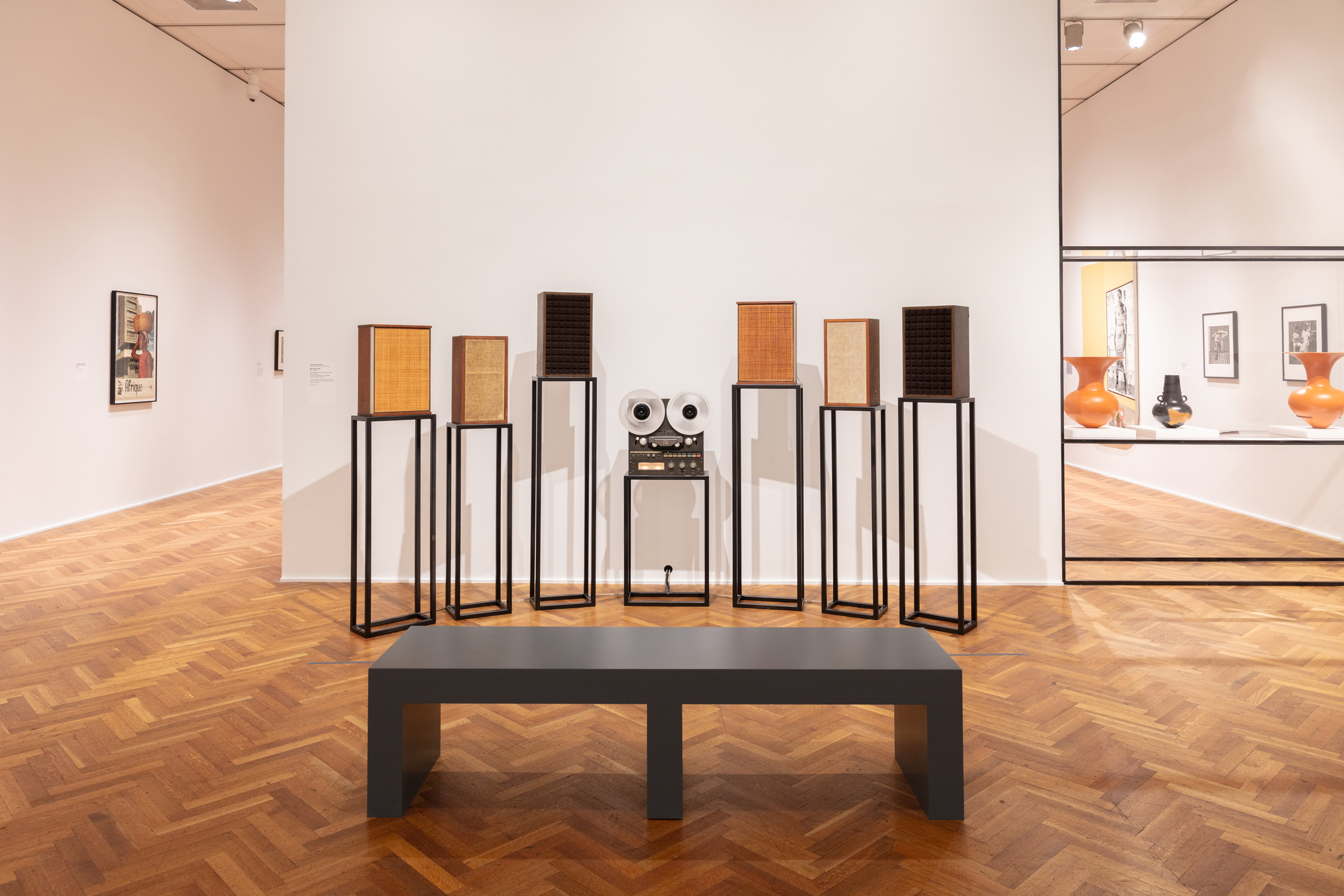 A modern gallery with a gray bench, vintage speakers on stands, a reel-to-reel tape recorder, framed artwork, and ceramics displayed on wooden flooring.